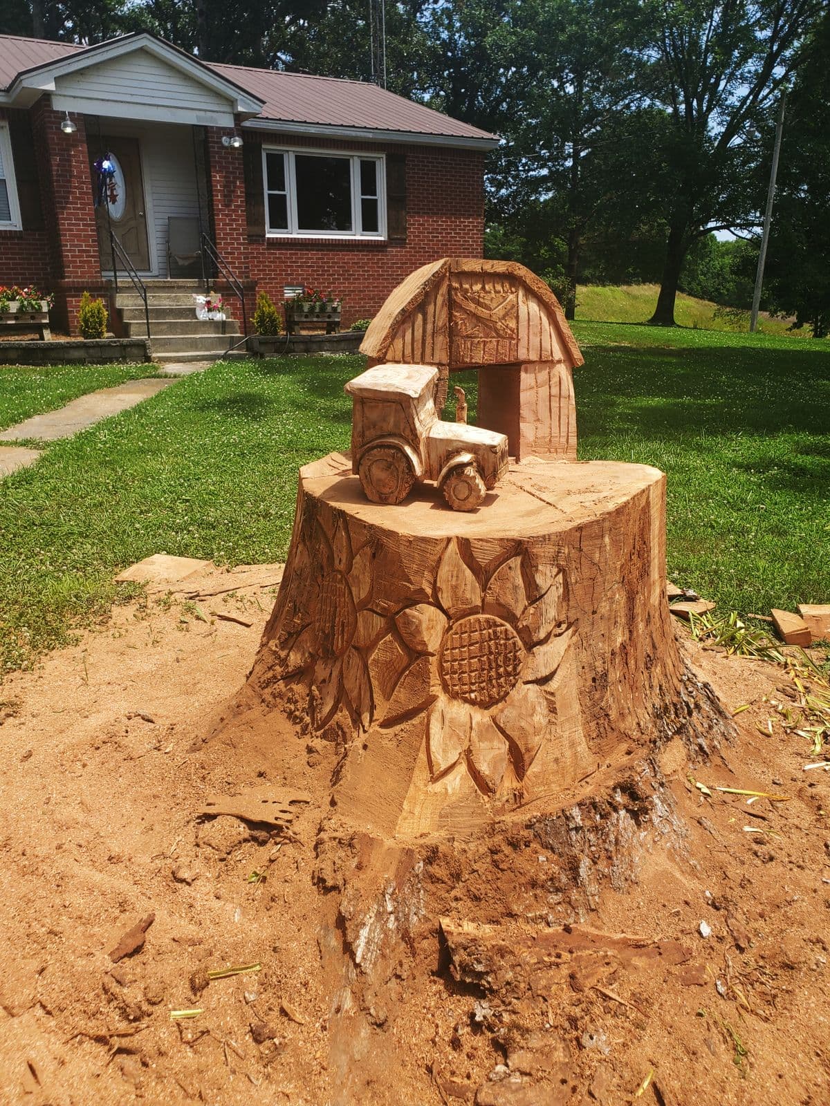 Front-yard tree stump carved into a barn and tractor with sunflowers around the base